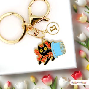 May include: Gold keyring with a black cat holding a blue pot with orange flowers. The keyring also has a gold circle charm with the letter "B" on it.