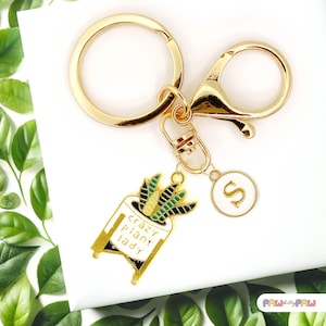 Könnte beinhalten: Goldener Schlüsselanhänger mit einer grünen Pflanze in einem weißen Topf mit dem Text "crazy plant lady" und einem goldenen Anhänger mit dem Buchstaben "S".