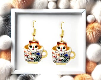 May include: A pair of gold-toned hook earrings featuring a colourful illustration of a cat peeking out from a floral teacup. The teacup design includes white, red, and blue floral patterns on a cream background with a gold handle and rim.