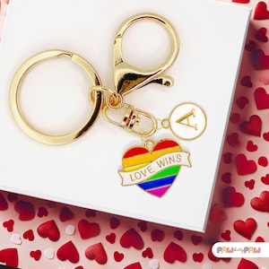 Könnte beinhalten: Goldfarbener Schlüsselanhänger mit einem herzförmigen Anhänger in Regenbogenfarben, mit dem Text "LOVE WINS" auf einem Banner. Enthält einen weißen runden Anhänger mit dem Buchstaben "A". Der Schlüsselanhänger liegt auf einer weißen Oberfläche mit roten herzförmigen Konfetti.
