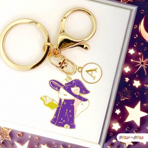 May include: Gold-tone keychain with a charm featuring a cartoon cat in a purple witch costume, holding a book. The charm has gold accents and a white circle with the letter "A". The keychain includes a clasp and a key ring.