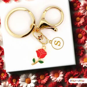 May include: Gold keyring with a red rose charm, a gold letter 'S' charm, and a gold split ring. The keyring is on a white surface surrounded by red and white flowers.