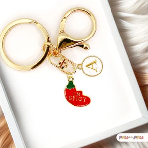 May include: Gold-coloured keychain with a lobster clasp, a round key ring, and a charm with the letter "A". A red chilli pepper charm with the words "I'M SPICY" is also attached. The keychain is on a white surface.