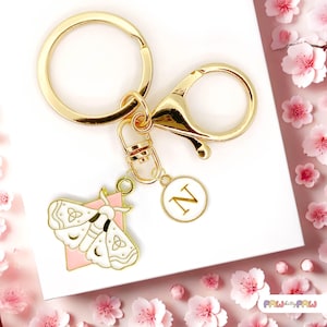 May include: Gold-coloured keyring with a lobster clasp, a round charm with the letter "N", and a moth-shaped charm with white and pink accents. The keyring is on a white surface with pink flowers.