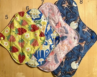 May include: Four reusable cloth menstrual pads labeled 5, 6, 7, and 8. Pad 5 is yellow with red strawberries. Pad 6 is blue with deer and mushrooms. Pad 7 is light pink with floral patterns. Pad 8 is dark blue with moths and moon motifs.