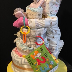 Puede incluir: Una cesta de regalo de baby shower de varios niveles con un body de beb&eacute;, un libro y otros art&iacute;culos para beb&eacute;s. La cesta est&aacute; decorada con un lazo rosa y una cinta blanca.