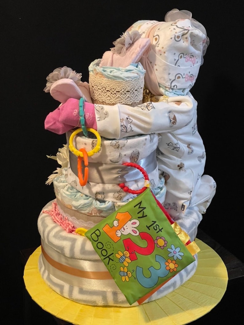 Puede incluir: Una cesta de regalo de baby shower de varios niveles con un body de beb&eacute;, un libro y otros art&iacute;culos para beb&eacute;s. La cesta est&aacute; decorada con un lazo rosa y una cinta blanca.
