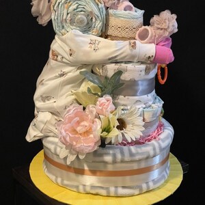 Puede incluir: Un regalo de baby shower de varios niveles hecho de pa&ntilde;ales, un body, un chupete y un sonajero. El regalo est&aacute; decorado con flores rosas y blancas, un lazo rosa y una cinta de encaje blanca.