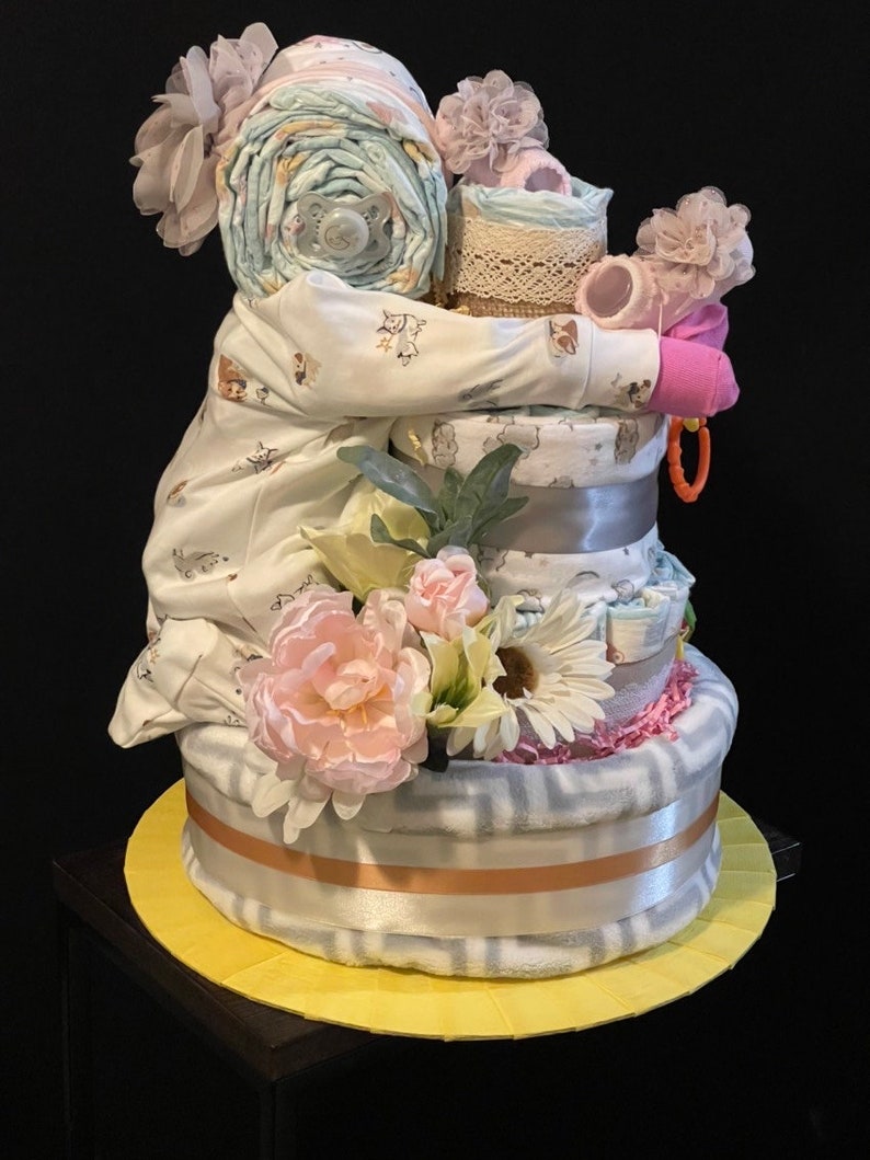 Puede incluir: Un regalo de baby shower de varios niveles hecho de pa&ntilde;ales, un body, un chupete y un sonajero. El regalo est&aacute; decorado con flores rosas y blancas, un lazo rosa y una cinta de encaje blanca.