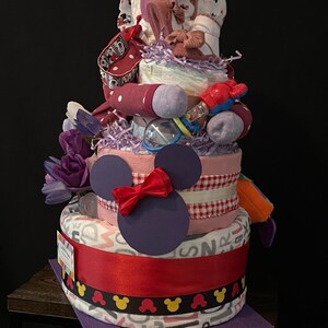 Puede incluir: Un pastel de pa&ntilde;ales de tres niveles decorado con un tema de Mickey Mouse en morado y rojo. El pastel est&aacute; adornado con un lazo rojo, una cabeza de Mickey Mouse morada y una cinta vichy blanca y roja. El nivel superior presenta un atuendo de beb&eacute; rosa y blanco con un dise&ntilde;o de Mickey Mouse.