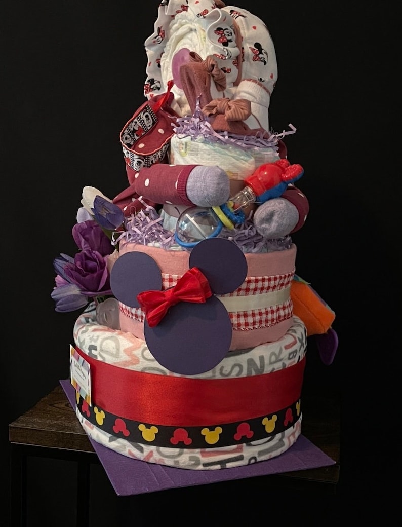 Puede incluir: Un pastel de pa&ntilde;ales de tres niveles decorado con un tema de Mickey Mouse en morado y rojo. El pastel est&aacute; adornado con un lazo rojo, una cabeza de Mickey Mouse morada y una cinta vichy blanca y roja. El nivel superior presenta un atuendo de beb&eacute; rosa y blanco con un dise&ntilde;o de Mickey Mouse.