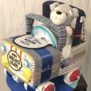 K&ouml;nnte beinhalten: Eine blaue und wei&szlig;e gestreifte Windeltorte in Form eines Autos mit einem grauen Teddyb&auml;ren, der auf dem Fahrersitz sitzt. Die Torte ist mit einem blauen Band, einem gelben und orangefarbenen gestreiften Band und einem gelben Schild mit der Aufschrift "baby hallock-kwon" verziert. Die Torte ist au&szlig;erdem mit einem blauen und wei&szlig;en gestreiften Stoff und einer blauen und wei&szlig;en gestreiften Schleife verziert. Die Torte steht auf einem blauen und wei&szlig;en gestreiften Sockel.