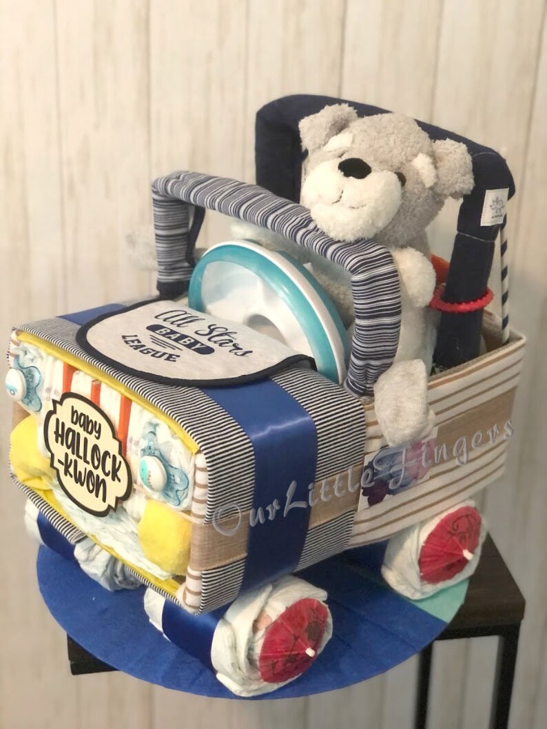 K&ouml;nnte beinhalten: Eine blaue und wei&szlig;e gestreifte Windeltorte in Form eines Autos mit einem grauen Teddyb&auml;ren, der auf dem Fahrersitz sitzt. Die Torte ist mit einem blauen Band, einem gelben und orangefarbenen gestreiften Band und einem gelben Schild mit der Aufschrift "baby hallock-kwon" verziert. Die Torte ist au&szlig;erdem mit einem blauen und wei&szlig;en gestreiften Stoff und einer blauen und wei&szlig;en gestreiften Schleife verziert. Die Torte steht auf einem blauen und wei&szlig;en gestreiften Sockel.