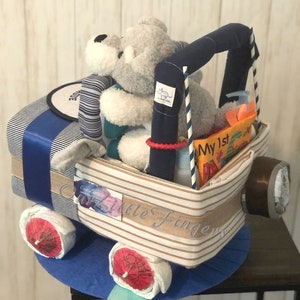 K&ouml;nnte beinhalten: Eine blaue und wei&szlig;e gestreifte Windeltorte in Form eines Spielzeuglastwagens mit einem grauen Stofftier auf dem Fahrersitz. Die Torte ist mit einem blauen Band und einem Schild verziert, auf dem "Mein 1. " steht.