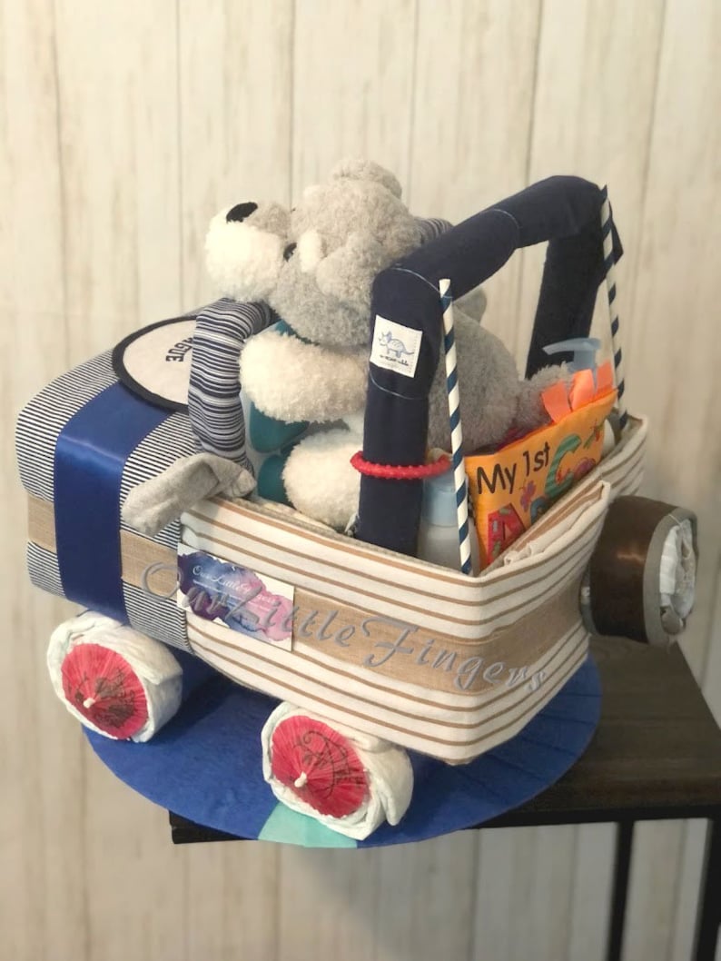 K&ouml;nnte beinhalten: Eine blaue und wei&szlig;e gestreifte Windeltorte in Form eines Spielzeuglastwagens mit einem grauen Stofftier auf dem Fahrersitz. Die Torte ist mit einem blauen Band und einem Schild verziert, auf dem "Mein 1. " steht.