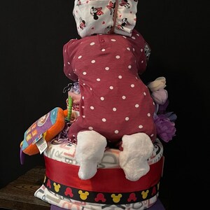 Puede incluir: Un pastel de pa&ntilde;ales con un mono de lunares rojos y blancos, una diadema blanca con un lazo rosa y una manta morada y blanca. El pastel est&aacute; decorado con una cinta roja y orejas de Mickey Mouse negras.