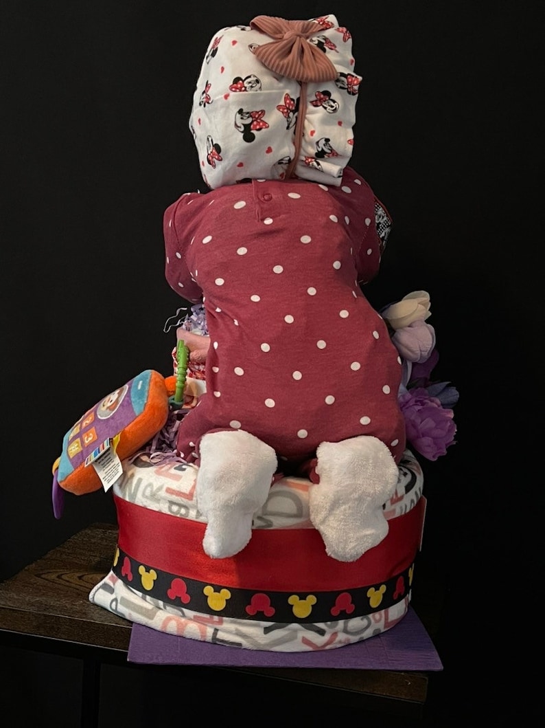 Puede incluir: Un pastel de pa&ntilde;ales con un mono de lunares rojos y blancos, una diadema blanca con un lazo rosa y una manta morada y blanca. El pastel est&aacute; decorado con una cinta roja y orejas de Mickey Mouse negras.