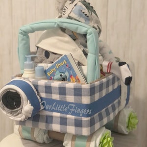 K&ouml;nnte beinhalten: Ein blauer und wei&szlig;er karierter Wickelkuchen in Form eines Lastwagens. Der LKW ist mit einem blauen Band verziert, auf dem "Our Little Fingers" steht. Der LKW ist gef&uuml;llt mit Babyartikeln, darunter ein Buch mit dem Titel "Baby Shark", ein Stofftier und eine Flasche Lotion.