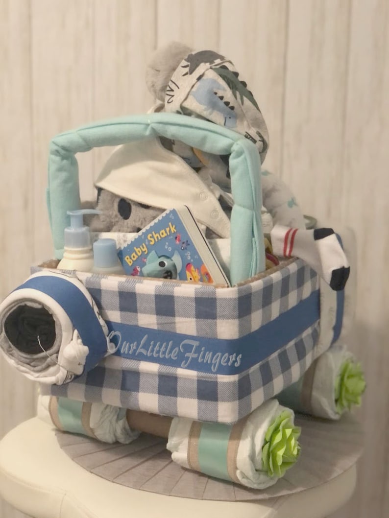 K&ouml;nnte beinhalten: Ein blauer und wei&szlig;er karierter Wickelkuchen in Form eines Lastwagens. Der LKW ist mit einem blauen Band verziert, auf dem "Our Little Fingers" steht. Der LKW ist gef&uuml;llt mit Babyartikeln, darunter ein Buch mit dem Titel "Baby Shark", ein Stofftier und eine Flasche Lotion.