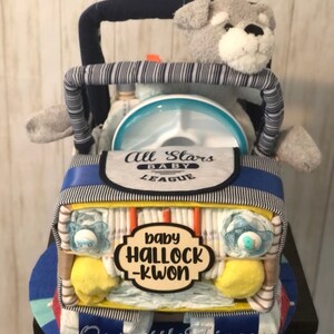 K&ouml;nnte beinhalten: Eine blaue und gelbe Windeltorte in Form eines Jeeps mit einem grauen Stoffhund auf dem Fahrersitz. Die Torte ist mit blauen und wei&szlig;en Streifen, einem blauen und wei&szlig;en L&auml;tzchen mit dem Text "All Stars Baby League" und einem Schild mit der Aufschrift "baby Halloch-Kwon" verziert.