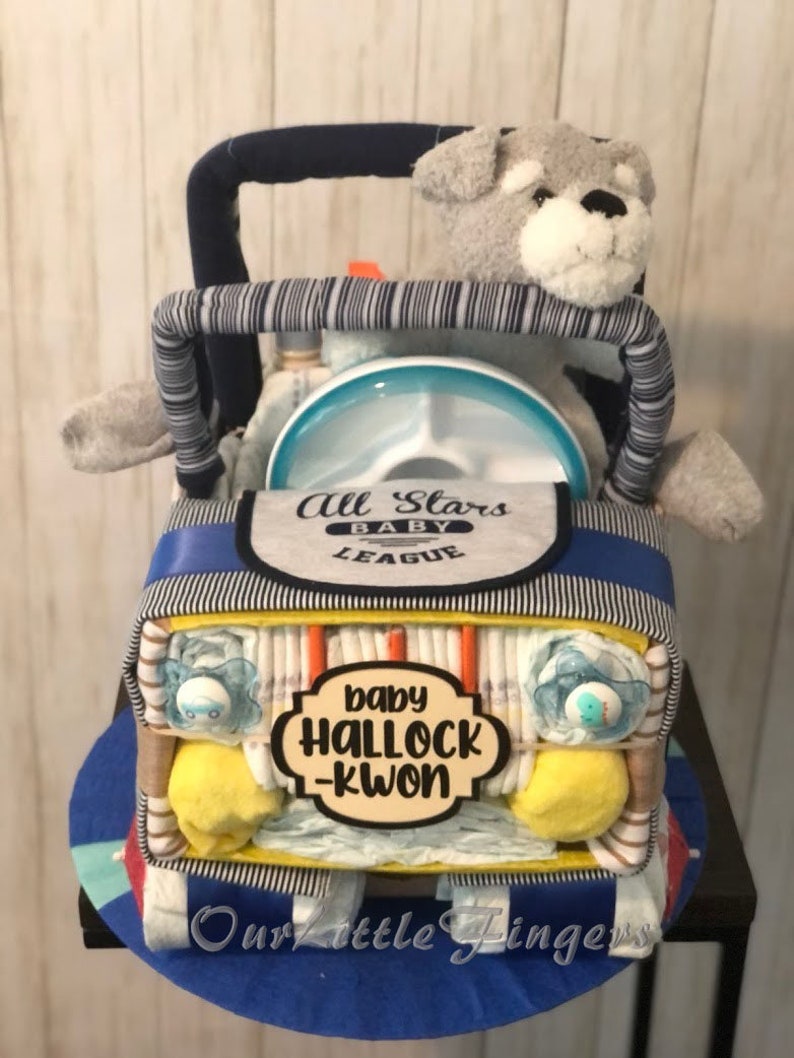 K&ouml;nnte beinhalten: Eine blaue und gelbe Windeltorte in Form eines Jeeps mit einem grauen Stoffhund auf dem Fahrersitz. Die Torte ist mit blauen und wei&szlig;en Streifen, einem blauen und wei&szlig;en L&auml;tzchen mit dem Text "All Stars Baby League" und einem Schild mit der Aufschrift "baby Halloch-Kwon" verziert.