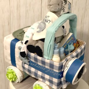 K&ouml;nnte beinhalten: Eine blaue und wei&szlig;e karierte Windeltorte in Form eines Spielzeuglastwagens. Der LKW hat ein blaues Band um sich und eine gr&uuml;ne Blume an jedem Rad. Der LKW ist gef&uuml;llt mit Babyartikeln, darunter ein Stofftier, ein Buch und Flaschen. Die Torte ist mit den Worten "Our Little Fingers" verziert.