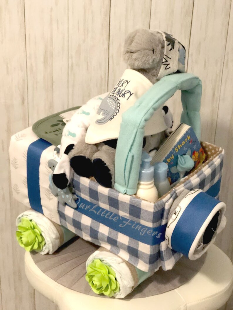 K&ouml;nnte beinhalten: Eine blaue und wei&szlig;e karierte Windeltorte in Form eines Spielzeuglastwagens. Der LKW hat ein blaues Band um sich und eine gr&uuml;ne Blume an jedem Rad. Der LKW ist gef&uuml;llt mit Babyartikeln, darunter ein Stofftier, ein Buch und Flaschen. Die Torte ist mit den Worten "Our Little Fingers" verziert.