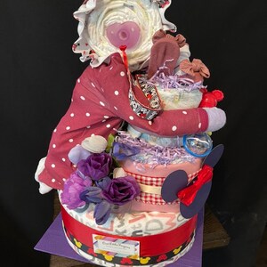 Puede incluir: Un pastel de pa&ntilde;ales de tres niveles decorado con una cinta de cuadros vichy rosa y blanca, flores moradas y un lazo rojo. El nivel superior presenta una mu&ntilde;eca beb&eacute; que lleva un atuendo de lunares rosa y blanco y un sombrero blanco. El pastel est&aacute; decorado con orejas de Mickey Mouse y el texto "One Little Fingers".
