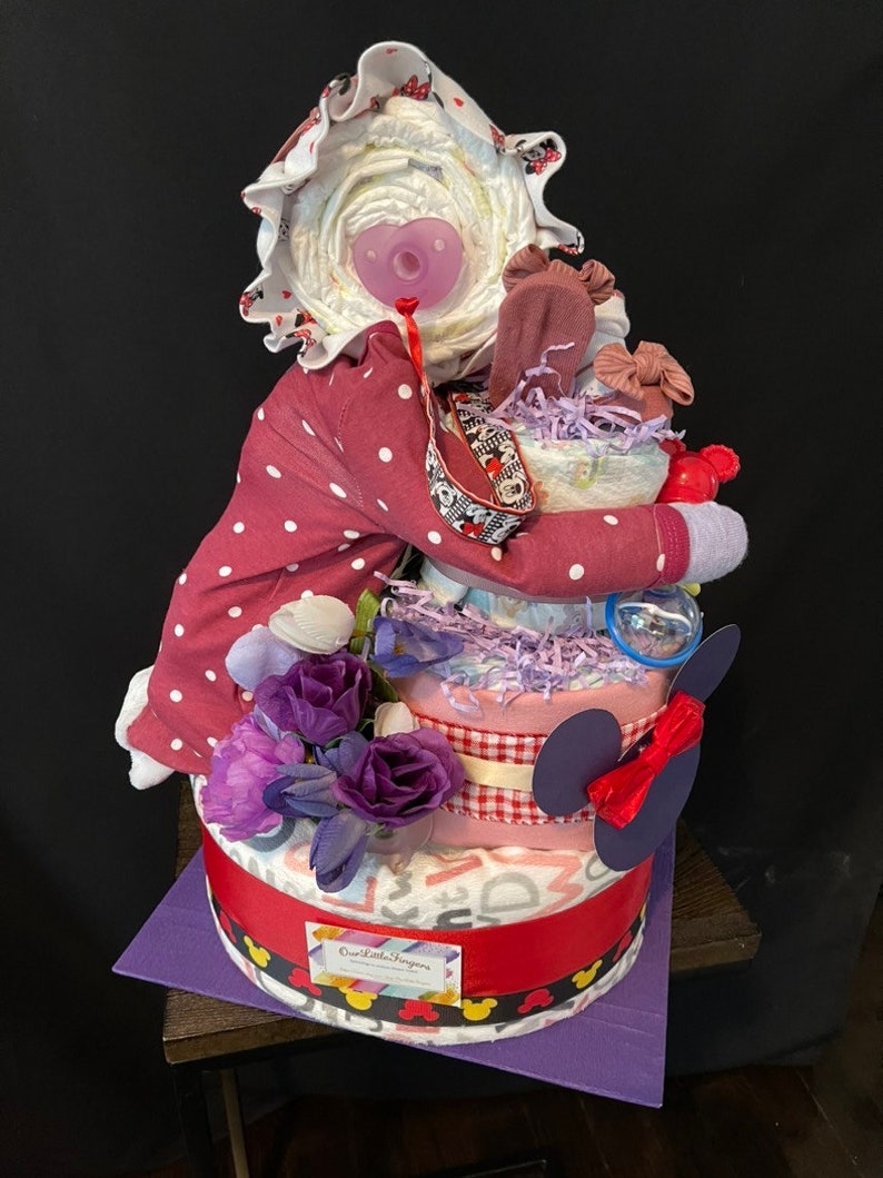 Puede incluir: Un pastel de pa&ntilde;ales de tres niveles decorado con una cinta de cuadros vichy rosa y blanca, flores moradas y un lazo rojo. El nivel superior presenta una mu&ntilde;eca beb&eacute; que lleva un atuendo de lunares rosa y blanco y un sombrero blanco. El pastel est&aacute; decorado con orejas de Mickey Mouse y el texto "One Little Fingers".