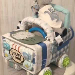 K&ouml;nnte beinhalten: Eine blaue und wei&szlig;e Windeltorte in Form eines Lastwagens mit einem grauen Dinosaurier-Pl&uuml;schtier oben drauf. Die Torte ist mit gr&uuml;nen und blauen Akzenten verziert und die Worte "Gavin A Paul" stehen an der Seite.