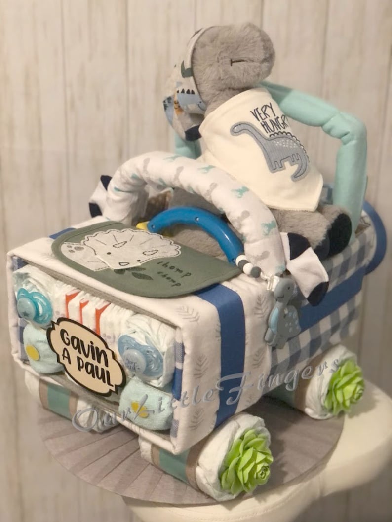 K&ouml;nnte beinhalten: Eine blaue und wei&szlig;e Windeltorte in Form eines Lastwagens mit einem grauen Dinosaurier-Pl&uuml;schtier oben drauf. Die Torte ist mit gr&uuml;nen und blauen Akzenten verziert und die Worte "Gavin A Paul" stehen an der Seite.