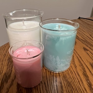 May include: Three glass candle jars with white, blue, and pink candles. The jars are graduated cylinders with markings in milliliters.