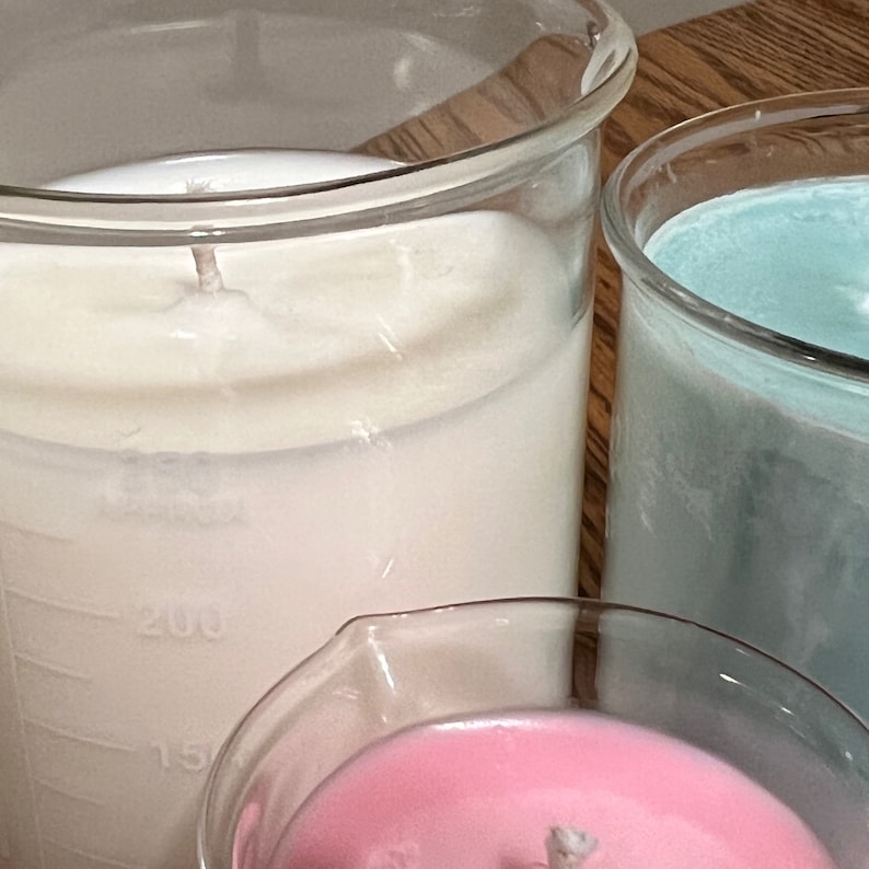 May include: Three glass containers with different colored wax inside. The containers are clear glass and have markings on the side. The wax is white, light blue, and pink.