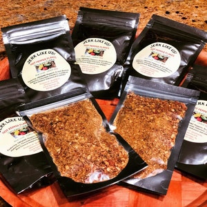 May include: Four small black pouches filled with a red and brown spice blend. The pouches are labeled "JERK LIKE US! HOT PEPPER FLAKES" and list the ingredients as "ORGANIC Peppers: Scotch Bonnet, Habanero,  Cayenne,  Red Chilli,  Habanada and GHOST".