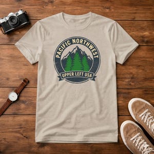 May include: A beige t-shirt with a circular graphic featuring the text "PACIFIC NORTHWEST" and "UPPER LEFT USA" surrounding a mountain and evergreen trees. The shirt is laid on a wooden surface with a watch, camera, and shoes.
