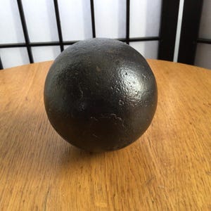 May include: A dark, spherical object with a textured surface, placed on a wooden table. The sphere is black and appears to be made of metal. The background features a window and a black lattice. The sphere's diameter is approximately 15 cm.