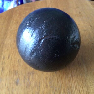 May include: A dark, spherical object with a textured surface, set on a wooden table. The sphere has a glossy appearance, possibly metal or painted. The item is likely a decorative piece or a paperweight.