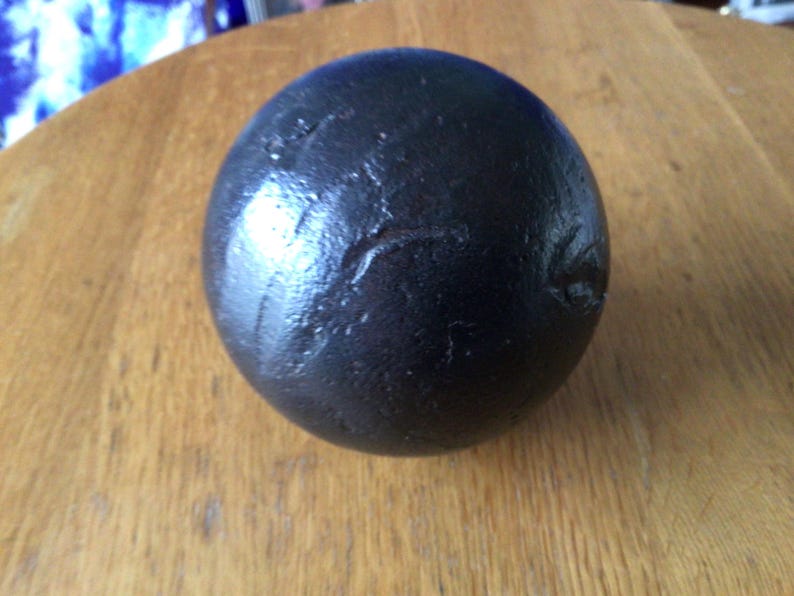 May include: A dark, spherical object with a textured surface, set on a wooden table. The sphere has a glossy appearance, possibly metal or painted. The item is likely a decorative piece or a paperweight.