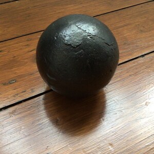 May include: A dark grey sphere with a textured surface, resting on a wooden surface. The wood has a warm brown tone and visible grain. The sphere's shape suggests it could be a decorative item or a historical artifact, possibly a cannonball.