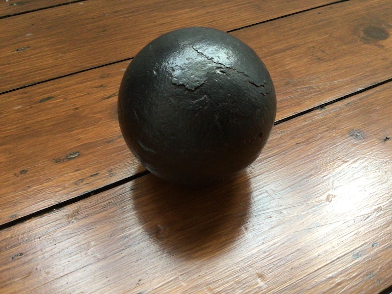 May include: A dark grey sphere with a textured surface, resting on a wooden surface. The wood has a warm brown tone and visible grain. The sphere's shape suggests it could be a decorative item or a historical artifact, possibly a cannonball.