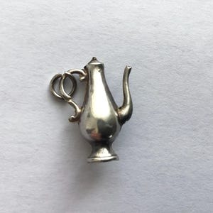 May include: A small, silver teapot charm with a curved spout and a looped handle. The charm has a detailed design and a small base. The teapot is likely made of metal and is set against a plain white background.
