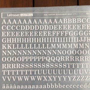 Puede incluir: Una hoja de rotulación instantánea Letraset con letras y números blancos de 42pt. Century Schoolbook Bold ATF. La hoja incluye letras mayúsculas y minúsculas, números y signos de puntuación. La hoja es gris.
