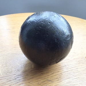 May include: A dark, spherical object with a textured surface, positioned on a light-coloured wooden table. The sphere is black and appears to be made of a dense material. The lighting creates highlights on the surface.