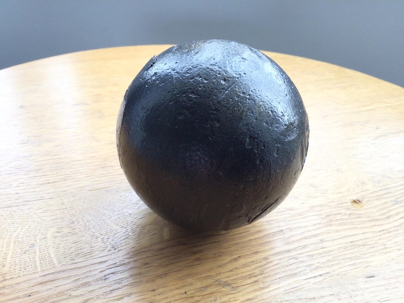 May include: A dark, spherical object with a textured surface, positioned on a light-coloured wooden table. The sphere is black and appears to be made of a dense material. The lighting creates highlights on the surface.