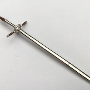 Może przedstawiać: Srebrny miniaturowy miecz z brązową rękojeścią i osłoną. Miecz ma szczegółową rękojeść i spiczaste ostrze. Projekt sugeruje motyw historyczny lub fantasy.