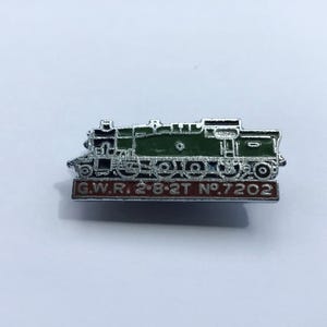 May include: A silver-toned metal enamel pin shaped like a steam locomotive. The train body is dark green with silver detailing. Below the train, a rectangular red base features the embossed silver text G.W.R. 2-8-2T N°.7202 in a clear font.