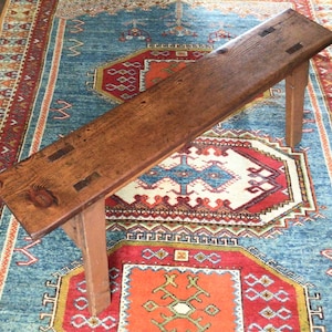 May include: A rectangular wooden bench with a dark brown stained finish, featuring a thick plank seat with visible wood grain, knots, and dark rectangular joinery details on the surface, supported by four sturdy, straight wooden legs.