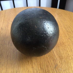 May include: A dark, spherical object with a textured surface. The object is resting on a light brown wooden surface. The sphere appears to be made of metal.