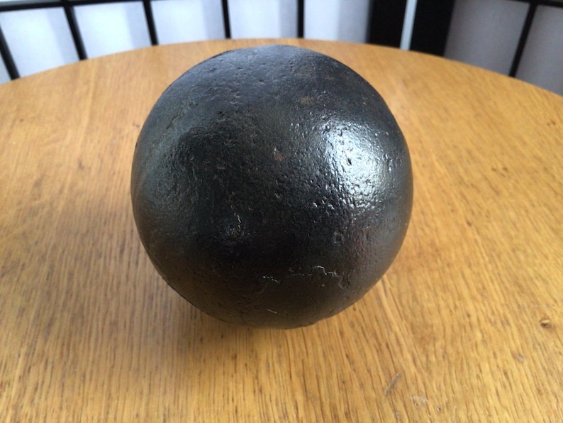 May include: A dark, spherical object with a textured surface. The object is resting on a light brown wooden surface. The sphere appears to be made of metal.