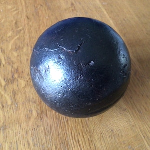 May include: A dark, spherical object with a textured surface and visible cracks. The object is a solid black colour and appears to be made of metal. It sits on a wooden surface.